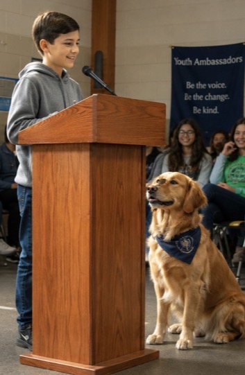 A middle-school ambassador speaking confidently at a small wooden podium with a working dog resting nearby.